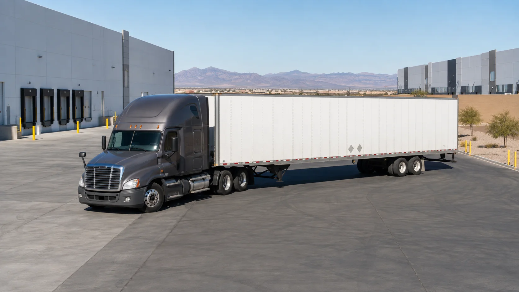 Graphite tractor and white trailer crossing a warehouse apron with desert mountains in the distance